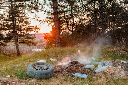 A car wheel next to a burning pile of leaves surrounded bu plastic bottles in the forest at sunrise.の写真素材