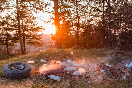 A car wheel next to a burning pile of leaves surrounded bu plastic bottles in the forest at sunrise.の写真素材