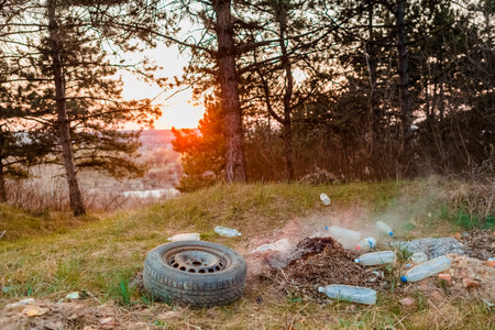A car wheel next to a burning pile of leaves surrounded bu plastic bottles in the forest at sunrise.の写真素材