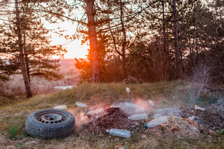 A car wheel next to a burning pile of leaves surrounded bu plastic bottles in the forest at sunrise.の写真素材