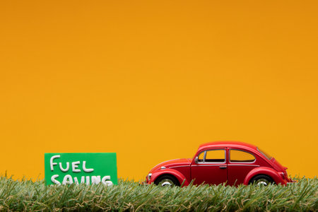Chisinau, Moldova - August 15th 2019: A red car figurine aligned to the right on grass next to a green sign with the words fuel saving on it, on orange background.のeditorial素材