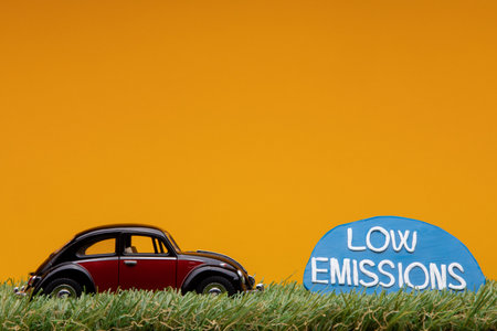 Chisinau, Moldova - August 15th 2019: A crimson car figurine aligned to the left on grass next to a pale blue sign with the words low emission on it, on orange background.のeditorial素材