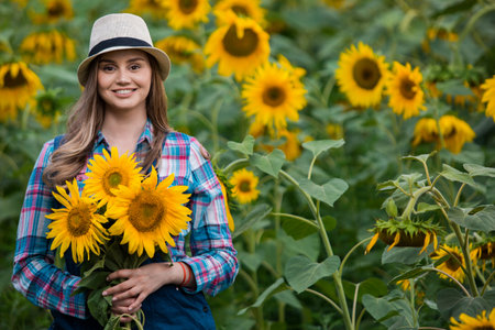 Gorgeous young female farmer standing in the middle of a green sunflower field, holding a bouquet of golden sunflowers during a sunny, beautiful day.の写真素材