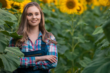 Gorgeous young female farmer standing in the middle of a green sunflower field with her hands crossed, during a sunny, beautiful day.の写真素材