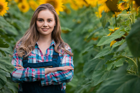 Gorgeous young female farmer standing in the middle of a green sunflower field with her hands crossed, during a sunny, beautiful day.の写真素材