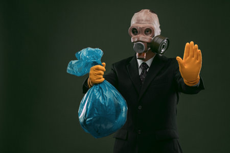 Portrait of a man wearing a dark business suit and an old gas mask holding a blue garbage bag and showing stop, isolated on dark green background.の写真素材