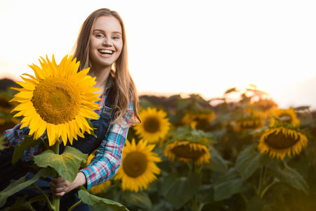 Gorgeous young female farmer standing in the middle of a green sunflower field next to a golden sunflower, during the early morning.の写真素材