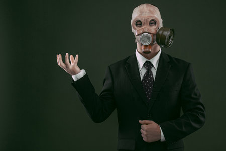 Portrait of a man wearing a dark business suit and an old gas mask holding his right hand up, isolated on dark green background.の写真素材