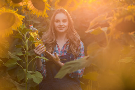 Gorgeous young female farmer standing in the middle of a green sunflower field holding a bottle of sunflower oil, during a majestic sunrise in the early morning.の写真素材