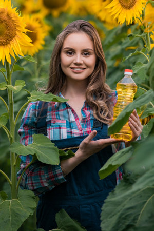 Gorgeous young female farmer standing in the middle of a green sunflower field holding a bottle of sunflower oil, during a sunny, beautiful day.の写真素材