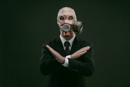 Portrait of a man wearing a dark business suit and an old gas mask with his hands x crossed, isolated on dark green background.の写真素材