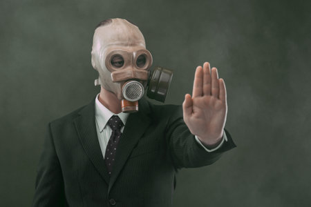 Portrait of a man wearing a dark business suit and an old gas mask showing his left palm, isolated on dark green background with fog.の写真素材