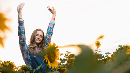 Gorgeous young female farmer standing in the middle of a green sunflower field with her hands up, during the early morning.の写真素材
