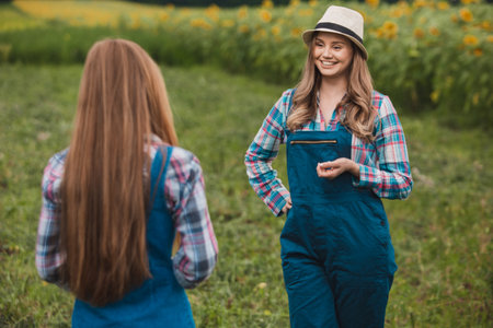 Two young, beautiful farmerettes having a discussion in the middle of a green blooming sunflower field.の写真素材