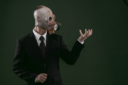 Portrait of a man wearing a dark business suit and an old gas mask holding his left hand up, isolated on dark green background.の写真素材