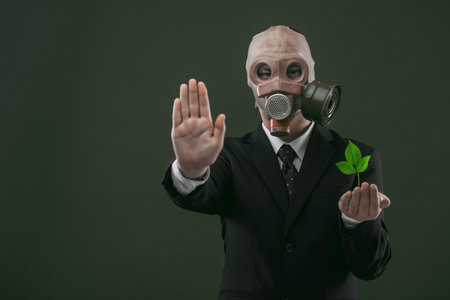 Portrait of a man wearing a dark business suit and an old gas mask while holding a green plant and showing stop, isolated on dark green background.の写真素材