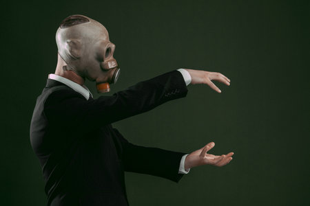 Portrait of a man wearing a dark business suit and an old gas mask holding his hands up, isolated on dark green background.の写真素材