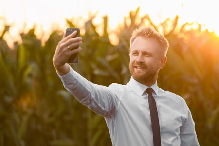 Handsome, blonde businessman taking a photo with a phone in the middle of a beautiful corn field, during sunrise.の写真素材