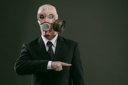 Portrait of a man wearing a dark business suit and an old gas mask pointing to the right, isolated on dark green background.の写真素材