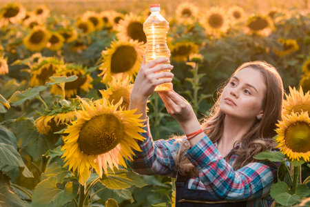 Gorgeous young female farmer standing in the middle of a green sunflower field holding a bottle of sunflower oil, during a majestic sunrise in the early morning.の写真素材