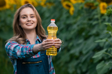 Gorgeous young female farmer standing in the middle of a green sunflower field holding a bottle of sunflower oil, during a sunny, beautiful day.の写真素材