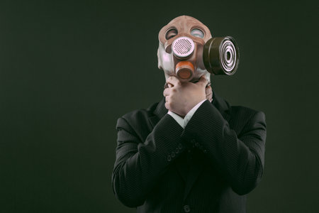 Portrait of a man wearing a dark business suit and an old gas mask while choking himself, isolated on dark green background.の写真素材
