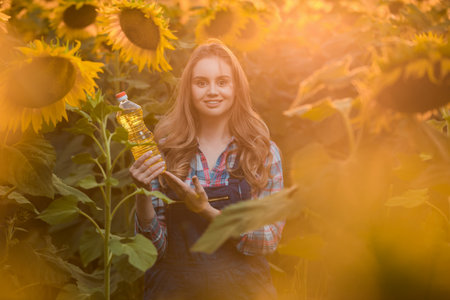 Gorgeous young female farmer standing in the middle of a green sunflower field holding a bottle of sunflower oil, during a majestic sunrise in the early morning.の写真素材