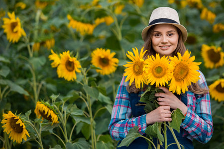 Gorgeous young female farmer standing in the middle of a green sunflower field, holding a bouquet of golden sunflowers during a sunny, beautiful day.の写真素材