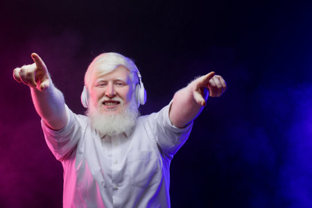 Portrait of an old albino grandfather pointing up, wearing headphones isolated on a beautiful, colorful background.の写真素材