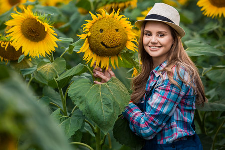 Gorgeous young female farmer standing in the middle of a green sunflower field near a sunflower with a smiley face, during a sunny, beautiful day.の写真素材