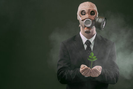 Portrait of a man wearing a dark business suit and an old gas mask while holding a green plant, isolated on dark green background with fog.の写真素材