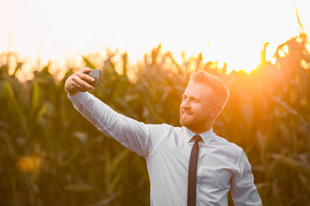 Handsome, blonde businessman taking a photo with a phone in the middle of a beautiful corn field, during sunrise.の写真素材