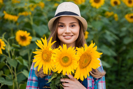 Gorgeous young female farmer standing in the middle of a green sunflower field, holding a bouquet of golden sunflowers during a sunny, beautiful day.の写真素材