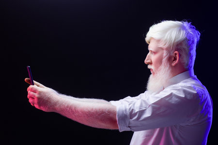 An old albino grandfather taking a photo with a modern phone, isolated on a beautiful, colorful background.の写真素材