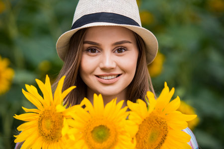 Gorgeous young female farmer standing in the middle of a green sunflower field, holding a bouquet of golden sunflowers during a sunny, beautiful day.の写真素材