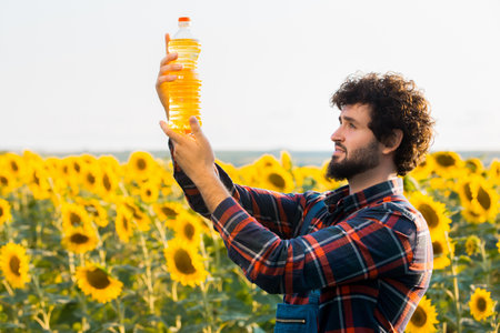 Handsome young farmer standing in the middle of a golden sunflower field holding up a sunflower oil bottle during a sunny, beautiful day.の写真素材
