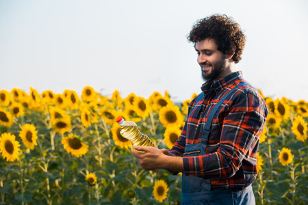 Handsome young farmer standing in the middle of a golden sunflower field smiling and examining a sunflower oil bottle during a sunny, beautiful day.の写真素材