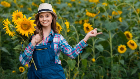Gorgeous young female farmer standing in the middle of a green sunflower field, holding a bouquet of golden sunflowers and pointing to her left during a sunny, beautiful day.の写真素材