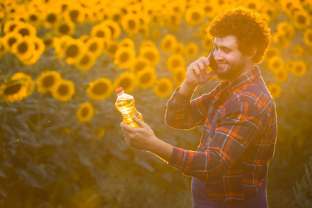 Handsome young farmer standing in the middle of a golden sunflower field smiling and talking on phone while holding up a sunflower oil bottle during a majestic sunrise.の写真素材