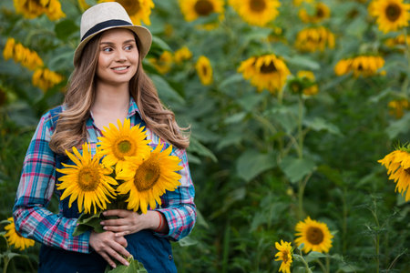 Gorgeous young female farmer standing in the middle of a green sunflower field, holding a bouquet of golden sunflowers during a sunny, beautiful day.の写真素材