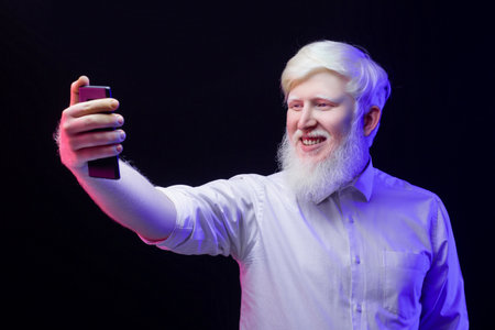 An old albino grandfather taking a photo with a modern phone, isolated on a beautiful, colorful background.の写真素材