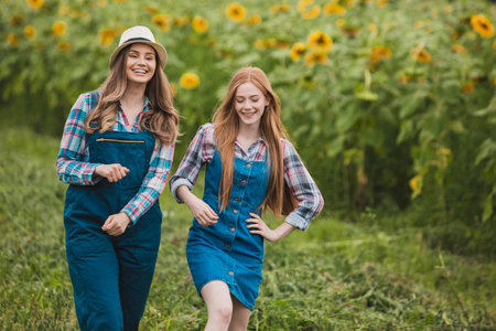 Two young, beautiful farmerettes walking in the middle of a green sunflower field, smiling.の写真素材