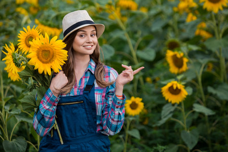 Gorgeous young female farmer standing in the middle of a green sunflower field, holding a bouquet of golden sunflowers and pointing to her left during a sunny, beautiful day.の写真素材