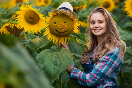 Gorgeous young female farmer standing in the middle of a green sunflower field near a sunflower with a smiley face, during a sunny, beautiful day.の写真素材
