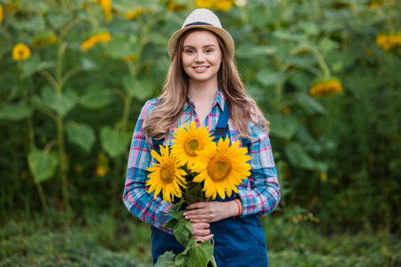 Gorgeous young female farmer standing in the middle of a green sunflower field, holding a bouquet of golden sunflowers during a sunny, beautiful day.の写真素材