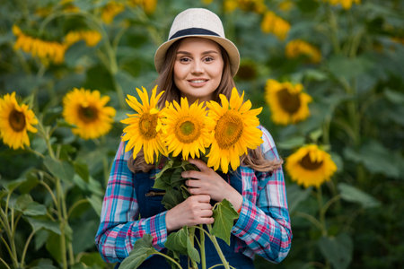Gorgeous young female farmer standing in the middle of a green sunflower field, holding a bouquet of golden sunflowers during a sunny, beautiful day.の写真素材