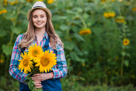 Gorgeous young female farmer standing in the middle of a green sunflower field, holding a bouquet of golden sunflowers during a sunny, beautiful day.の写真素材