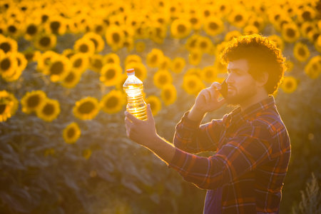 Handsome young farmer standing in the middle of a golden sunflower field talking on phone while holding up a sunflower oil bottle during a majestic sunrise.の写真素材