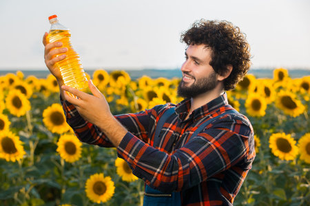 Handsome young farmer standing in the middle of a golden sunflower field smiling and holding up a sunflower oil bottle during a sunny, beautiful day.の写真素材