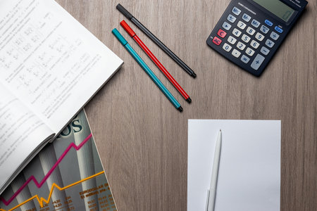 overhead view of a table with desk or office items, with open books, pens and a calculator.の写真素材